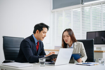 Two Asian business professionals in formal suits discussing a project at modern office desk with laptop and documents.