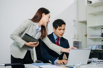 Young Asian businessman and colleague using laptop and tablet while discussing project in modern...