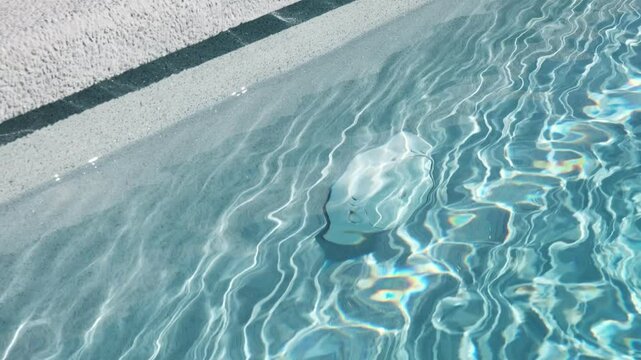 Underwater section of a swimming pool with clear blue water and an integrated plastic circulation system element. Bright sunlight reflections create a vibrant light pattern on the water

