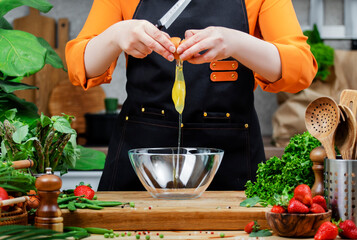 Chef woman preparing healthy dish, cracking egg into bowl while surrounded by fresh vegetables and kitchen utensils.
