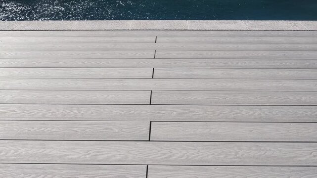 Light gray composite deck boards with wood texture by the pool edge. Modern surface creates a neat and aesthetic relaxation area by the water, reflecting sunlight beautifully
