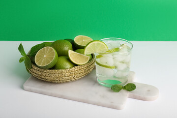 Glass of fresh Mojito cocktail and wicker bowl with limes on white table against green background