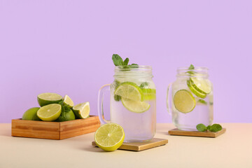 Mason jars of fresh Mojito cocktail with mint and limes on beige table against lilac background