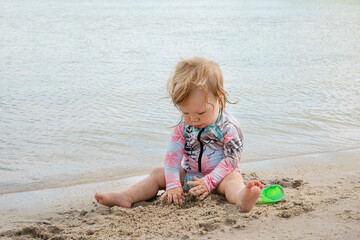 Adorable toddler girl sitting at the edge of the sea on sandy beach, wearing colorful sun protective swimsuit, playing with wet sand, exploring shoreline, carefree childhood summer vacation moment out