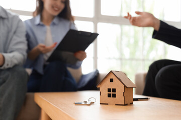 House figure and key on table against couple signing agreement with real estate agent in room,...