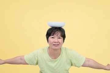 middle age asian woman balancing empty bowl on head, playful pose on studio background