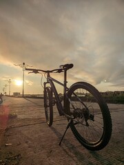 A bicycle sits parked on a concrete path during a golden sunset, casting long shadows around it.
