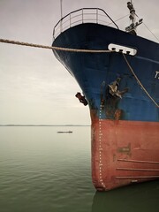 Bow of a blue and red ship with anchor and a distant boat on calm, muted colored water.