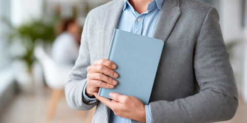A person in a gray blazer holds a light blue book or notebook close to their chest in an indoor setting with blurred background.