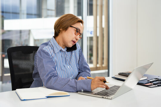 mature middle aged business woman, 40s professional lady executive manager talking on the phone making business call on cellphone at work in office using laptop computer. - Powered by Adobe