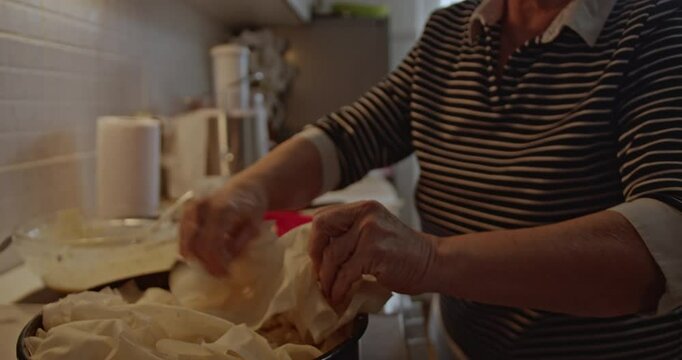 An elderly woman is carefully preparing a traditional pastry in her cozy home kitchen. She layers filo dough and adds a savory filling.