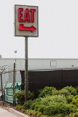 Vintage red neon “EAT” arrow sign on corrugated metal panel above industrial fence and greenery...