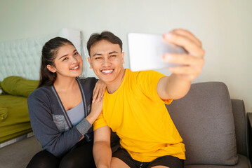 Indonesian southeast asian couple relaxing while taking a selfie or video call with a cellphone on the sofa at home