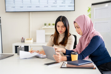 Concentrated young woman in hijab sitting with asian colleague at table, looking at lablet screen, explaining new company job. Focused team leader training female intern.