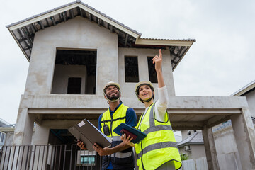 Two construction workers are standing in front of a house. One of them is pointing at the house