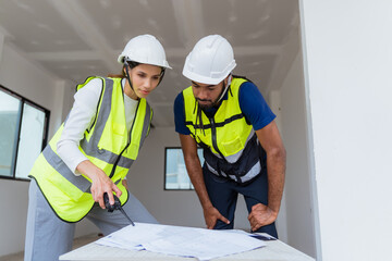Two men are looking at a blueprint. One of them is wearing a yellow vest. The other man is wearing...