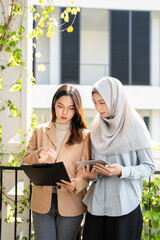 Two businesswomen, one wearing a hijab, standing in ffice building corridoor, smiling and talking...