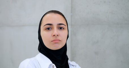 Professional muslim female healthcare worker wearing hijab and white coat, standing against concrete wall, displaying confident facial expressions while facing camera