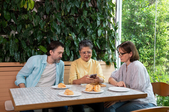 Middle-aged Asian woman using smartphone while sitting with middle-aged Caucasian man and woman at table with pastries, concept of multicultural friendship, sharing, and digital communication