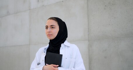 Young muslim woman in hijab leaning against concrete wall, holding book and gazing thoughtfully, reflecting on academic goals and professional aspirations
