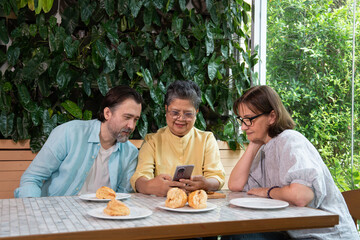 Middle-aged Asian woman showing smartphone to middle-aged Caucasian man and woman while sitting together at table with pastries, concept of cultural exchange, digital connection and shared learning