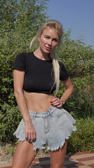 Blonde tanned Caucasian woman with navel piercing wears denim shorts and black cropped top in a park
