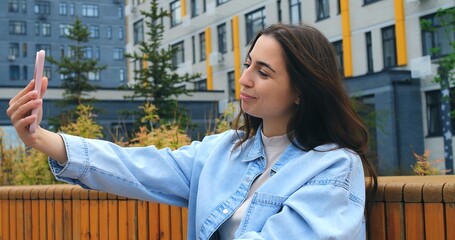 Outdoors, a young woman with long brown hair, wearing a light denim jacket, holds her smartphone and speaks into it, likely recording a voice message or using a voice assistant