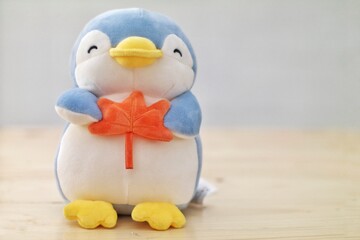 A plush penguin toy holding an orange leaf, sitting on a light wooden surface with a soft background