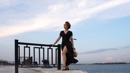 Confident woman in black dress overlooks calm seaside horizon. A lady with beautiful long legs...
