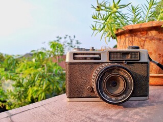 Vintage 50mm camera set on a table outdoors with greenery and a clay pot in the background. Warm...