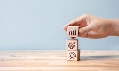 A hand stacking wooden blocks with business icons representing growth, target achievement, and process optimization, symbolizing strategic planning and development.