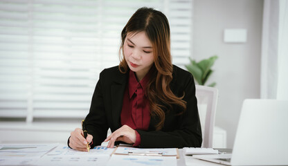 A young Asian businesswoman in a black blazer and red shirt calculates numbers on a white...