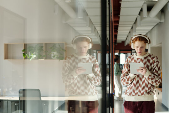 Young Caucasian man with headphones standing in modern coworking space using digital tablet, reflection visible in glass wall