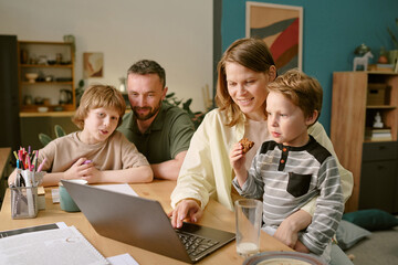 Caucasian middle aged woman sitting at desk with two Caucasian boys and Caucasian middle aged man, using laptop together, younger boy eating cookie, working from home