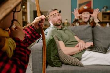Caucasian middle aged man working from home on laptop sitting on sofa while two children playing in background, one child aiming toy bow and arrow, other child making playful gesture