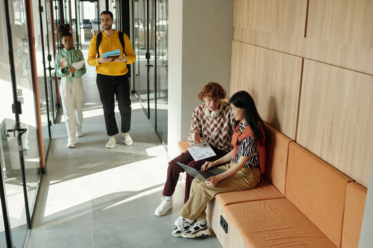 Young Caucasian man and Asian woman sitting on bench collaborating with laptop while Black colleagues walking in modern coworking hallway - Powered by Adobe