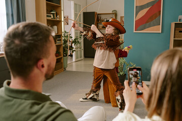 Caucasian child wearing cowboy costume performing with toy bow and arrow in living room, while young Caucasian man and woman watching and recording with smartphone during work from home
