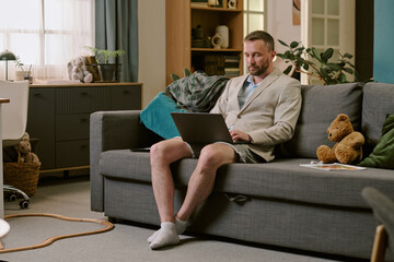 Middle aged Caucasian man sitting on sofa using laptop for remote work wearing business jacket and...