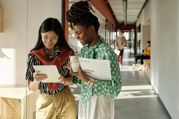 Young adult Asian woman holding digital tablet standing next to Black woman with dreadlocks holding...