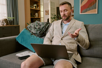 Caucasian middle aged man sitting on sofa using laptop for remote work, gesturing with hand during...