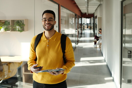 Biracial man with beard wearing yellow sweater and glasses standing in modern coworking space holding smartphone and digital tablet smiling at camera