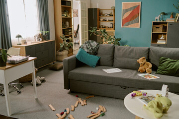 Modern living room with scattered wooden toy train tracks, plush teddy bear on gray sofa, children drawings on table, home office desk in background suggesting work from home setting