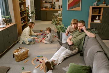 Caucasian middle aged man multitasking with laptop and smartphone on sofa, while Caucasian boy...