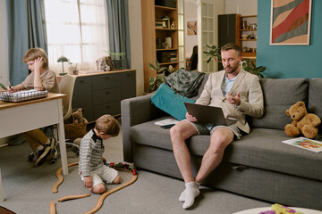 Caucasian middle aged man sitting on sofa using laptop for remote work wearing blazer and shorts,...