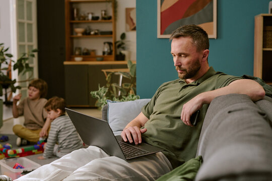 Middle aged Caucasian man working on laptop, sitting on sofa in living room while two young Caucasian boys playing with toys on floor in background during work from home scenario