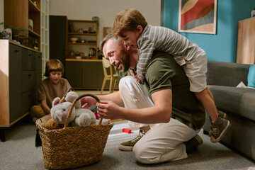 Middle aged Caucasian man kneeling on floor playing with stuffed animals in basket, while young...