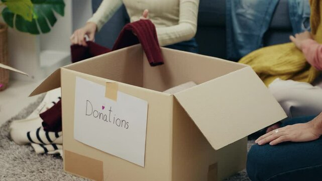 Close-up view of people placing folded clothes into a donation box. The teamwork and support represent a meaningful act of kindness in a home setting.