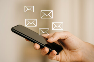 Hand holding black smartphone with white email icons floating above it beige background mobile messaging communication concept