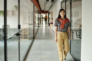 Young adult Asian woman walking through modern coworking office hallway holding laptop, people...