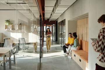 Asian woman walking through modern coworking space while Black woman and man sitting on bench talking, Caucasian man standing with tablet